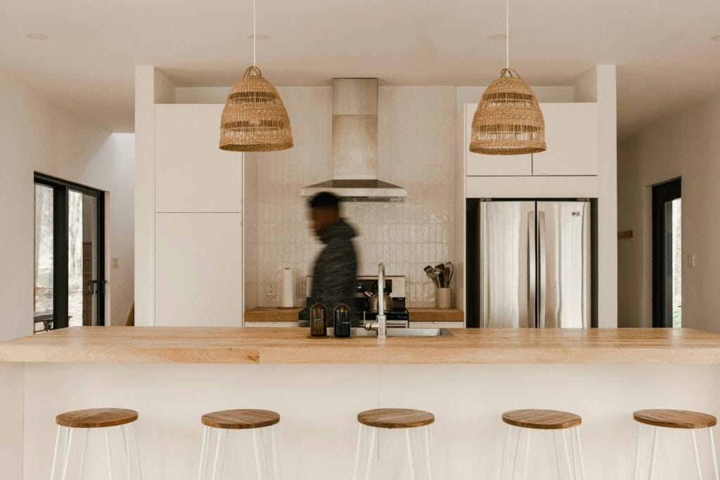 Modern kitchen with wooden accents, a person walking by, island with stools, pendant lights, LG GF-L700MWH stainless steel fridge, and range hood.