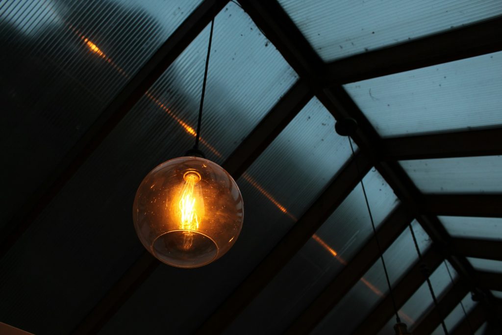 A round incandescent light bulb hangs from the ceiling, glowing warmly against the backdrop of a dark, slanted glass roof.